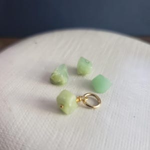 May include: A collection of light green gemstones, some rough-cut and others polished, scattered on a white surface. One gemstone is attached to a small gold-colored ring. The background is a gradient of white to dark blue.