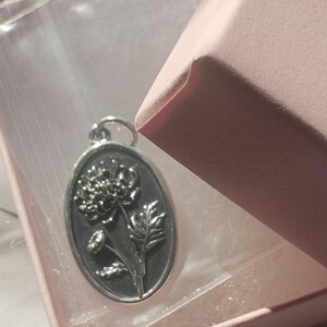 May include: Oval-shaped silver pendant featuring a detailed floral design. The pendant is suspended from a small loop and is presented inside a pink gift box. The floral design includes a large flower, leaves, and smaller buds.