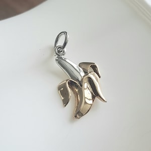 May include: A silver and gold banana charm pendant. The banana is made of two metals, with the top half being silver and the bottom half being gold.