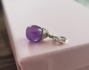 May include: A faceted, teardrop-shaped amethyst gemstone pendant. The purple stone is wrapped in silver-coloured wire and attached to a small silver ring. The pendant is displayed on a light pink surface.