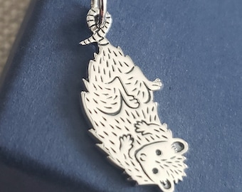 May include: A silver charm in the shape of a curled up hedgehog. The charm is detailed with a textured body and facial features.