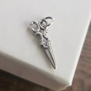 May include: A silver charm in the shape of a pair of scissors. The scissors have a detailed design and a loop at the top for attaching to a bracelet or necklace.