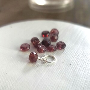 May include: A close-up of a small, faceted, dark red gemstone charm with a silver jump ring. The charm is surrounded by several other faceted, dark red gemstones.