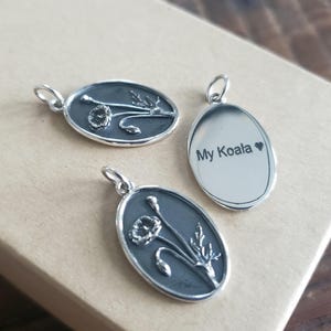 Puede incluir: Tres abalorios ovalados de plata. Un abalorio presenta un diseño de amapola, otro abalorio presenta un diseño de amapola y el tercer abalorio tiene el texto "My Koala" grabado en él.