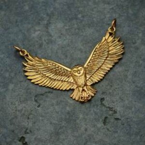 May include: A gold-toned pendant necklace featuring a detailed, three-dimensional owl with its wings spread wide. The owl is facing forward with its eyes open and its talons visible.