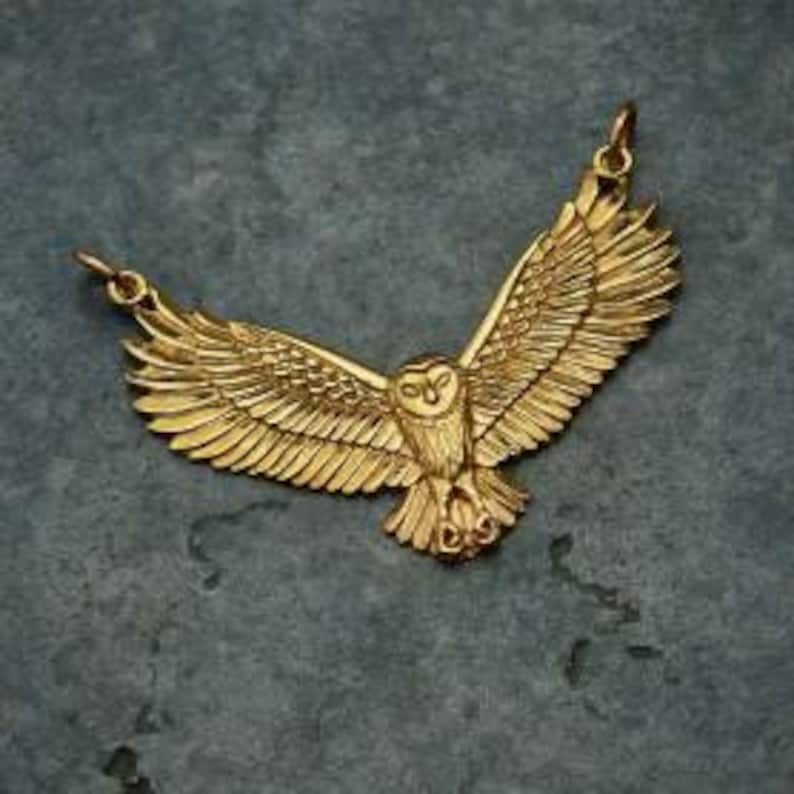 May include: A gold-toned pendant necklace featuring a detailed, three-dimensional owl with its wings spread wide. The owl is facing forward with its eyes open and its talons visible.
