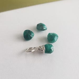 May include: A collection of faceted, emerald-green gemstones. One gemstone is wire-wrapped with a silver-colored bail, suitable for a pendant. Three loose gemstones are scattered nearby. The background is white with a hint of a gold ribbon.