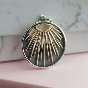 以下が含まれることがあります： 太陽の光が放射状に広がるデザインの、銀と金の丸いペンダント。ペンダントは黒色の背景に、金色の太陽から放射状に広がる金色の光線がある。