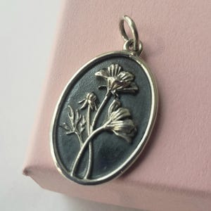 May include: Oval silver pendant with a floral design. The pendant features a dark background with a raised relief of flowers and leaves. The pendant has a silver border and a loop for a chain.