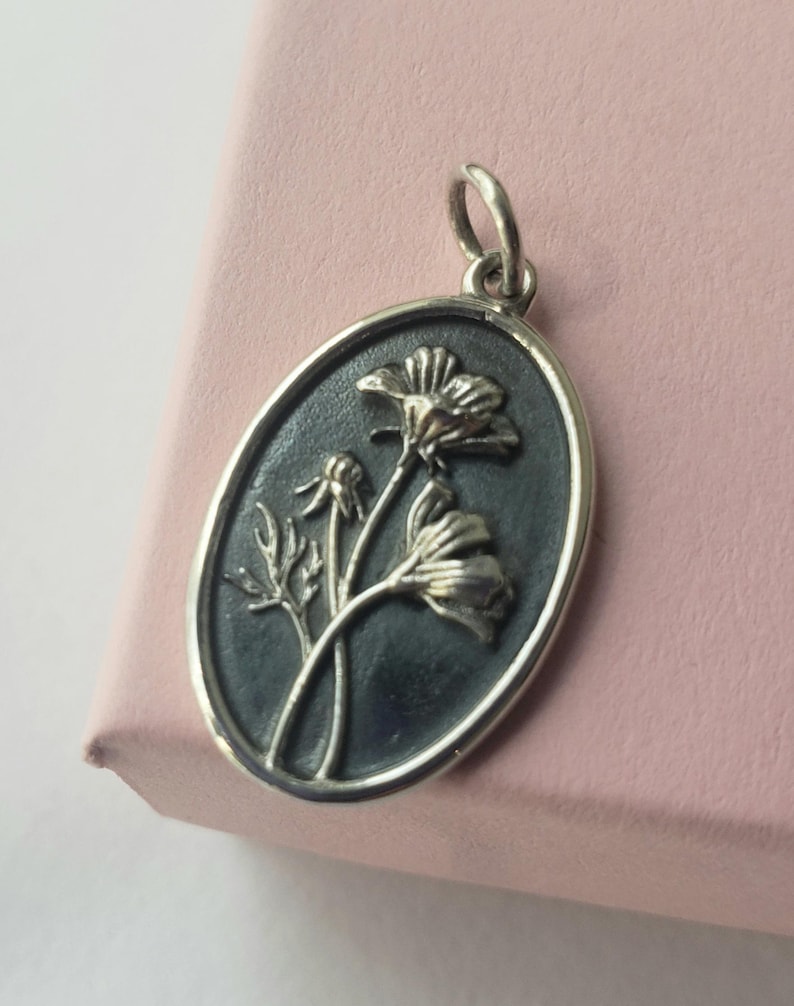 May include: Oval silver pendant with a floral design. The pendant features a dark background with a raised relief of flowers and leaves. The pendant has a silver border and a loop for a chain.