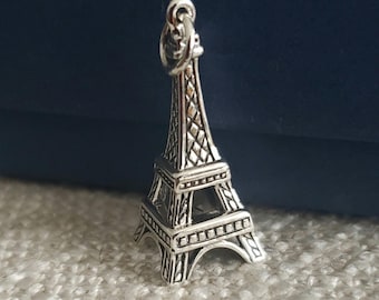 May include: A silver-colored Eiffel Tower charm with intricate detailing. The charm features a loop at the top for attachment and is set against a neutral background. The design captures the iconic structure's architectural elements.