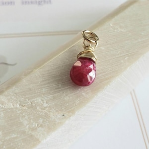 Puede incluir: Un colgante de piedra preciosa de rubí facetado en forma de lágrima. El rubí es de color rojo intenso y está envuelto en alambre dorado. El colgante tiene un lazo dorado en la parte superior para colgar. Fondo claro.