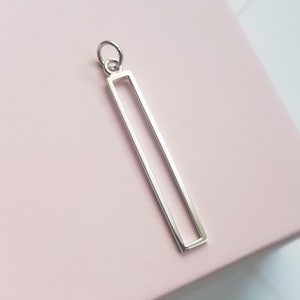 May include: A silver rectangular pendant with a small loop at the top. The pendant has a simple, open-frame design, with clean lines and a polished finish. The pendant is shown against a soft pink background.