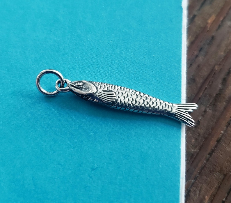 May include: A silver-colored fish pendant with detailed scales, fins, and a loop for a chain. The pendant is a small, detailed representation of a fish, perfect for jewelry or crafting projects.
