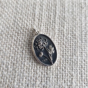 May include: Oval silver pendant featuring a detailed floral design. The pendant showcases a dark background with a raised, stylized flower and leaves. The pendant is suspended from a small silver loop.