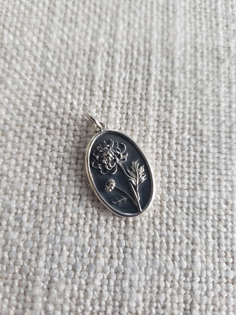 May include: Oval silver pendant featuring a detailed floral design. The pendant showcases a dark background with a raised, stylized flower and leaves. The pendant is suspended from a small silver loop.