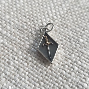 Puede incluir: Un amuleto de plata en forma de diamante con una daga dorada en su interior. El amuleto tiene un bucle en la parte superior para sujetarlo a un collar o pulsera.