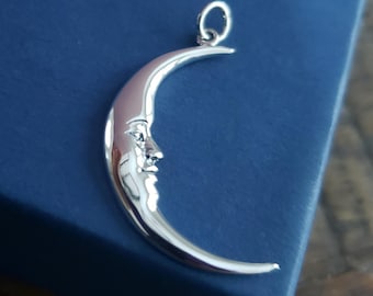 May include: A silver crescent moon pendant with a detailed face design. The pendant is suspended from a small loop at the top. The moon is set against a dark blue background, creating a striking contrast.