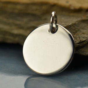 May include: A shiny, silver-colored, round pendant with a small loop at the top. The pendant is smooth and reflective, with a polished surface. The jewelry is likely made of sterling silver or a similar metal.