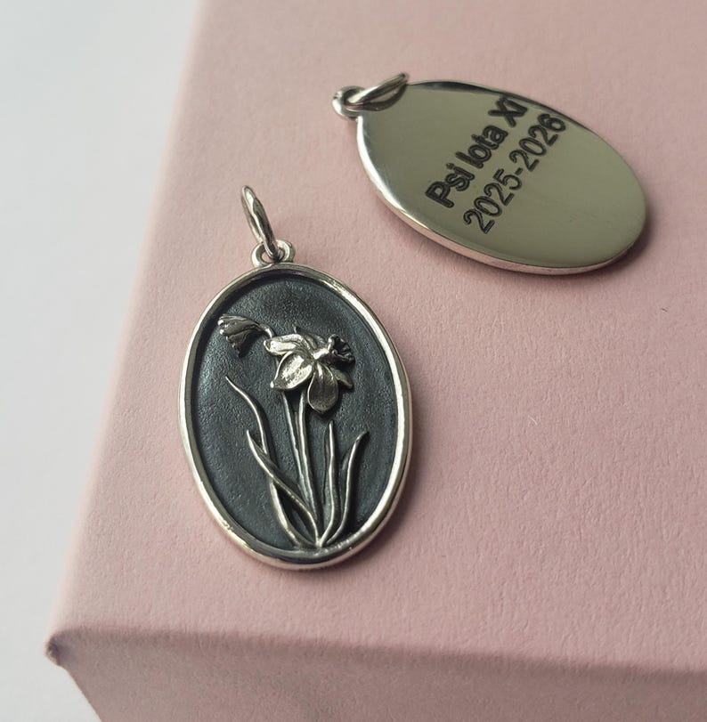 May include: Two silver pendants. One oval pendant features a daffodil design with a dark background. The other oval pendant has the text "Psi Iota Xi 2025-2026" engraved on it. Both pendants have a loop for a chain.