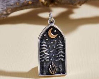 Puede incluir: Colgante de plata con una imagen en blanco y negro de dos pinos, una luna creciente y estrellas sobre un fondo oscuro. La luna está acentuada en oro.