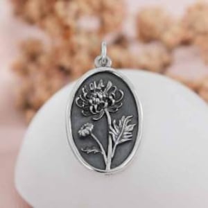 May include: A silver-colored pendant with an oval shape. The pendant features a detailed floral design, including a large flower and a smaller bud, set against a dark background. The pendant is suspended from a small loop at the top.
