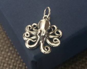 Puede incluir: Un colgante de pulpo plateado con tentáculos detallados y un lazo para una cadena. El colgante se encuentra sobre un fondo azul oscuro, mostrando su intrincado diseño y brillo metálico. Este dije de temática náutica es adecuado para joyería.