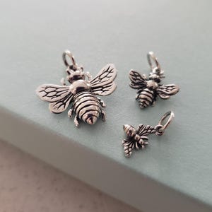 May include: Three silver bee charms of varying sizes. The largest charm is a detailed bee with wings outstretched. The two smaller charms are simpler bee designs.