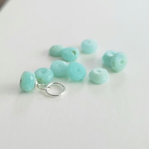 May include: A collection of small, faceted, light turquoise beads with a smooth, rounded shape. One bead is attached to a circular silver-toned metal jump ring, while the other eight beads are scattered loosely around it on a plain surface.