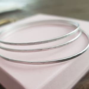 May include: Three silver hammered bangle bracelets stacked on top of each other on a pink surface.