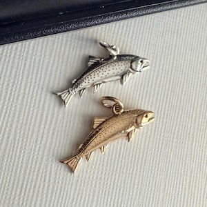 May include: Two fish-shaped charms, one in a polished silver tone and one in a warm gold tone. Each charm features detailed scales, fins, and a small circular jump ring at the top for attachment. The fish are depicted in a swimming position.