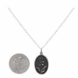 May include: Silver-toned necklace featuring an oval pendant with a lily of the valley design. The pendant has a dark background and a detailed floral motif. The chain is delicate, and a dime is included for size reference.