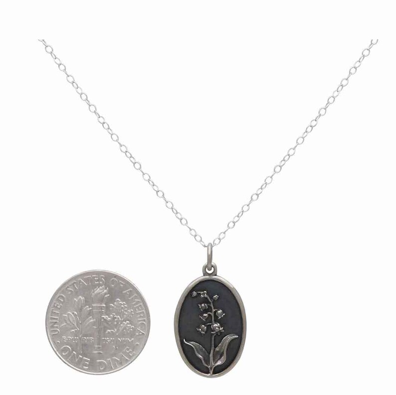 May include: Silver-toned necklace featuring an oval pendant with a lily of the valley design. The pendant has a dark background and a detailed floral motif. The chain is delicate, and a dime is included for size reference.