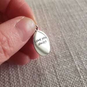 May include: A small, oval-shaped silver pendant with the engraved words "Love you always." The pendant has a loop at the top for a chain. The background is a textured, neutral-colored fabric.