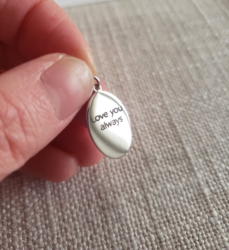 May include: A small, oval-shaped silver pendant with the engraved words "Love you always." The pendant has a loop at the top for a chain. The background is a textured, neutral-colored fabric.
