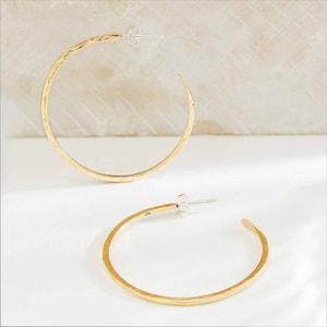 May include: Two gold-coloured hoop earrings with a hammered texture. The earrings have a simple, elegant design with a post closure. The earrings are displayed on a white surface.