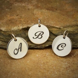 May include: Three silver-coloured round pendants, each with a different black cursive letter: A, B, and C. Each pendant has a small loop at the top for hanging. The pendants are displayed on a textured, brown surface.