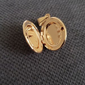 May include: A gold-toned oval locket with a hinged lid. The locket is open, revealing a blank space inside. The locket has a small loop at the top for attaching a chain.