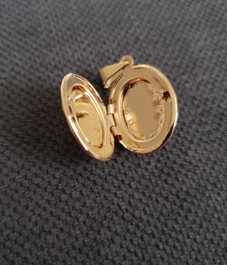 May include: A gold-toned oval locket with a hinged lid. The locket is open, revealing a blank space inside. The locket has a small loop at the top for attaching a chain.