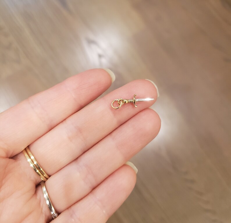 May include: A gold charm in the shape of a silver dagger with a gold loop for attaching to a necklace or bracelet.