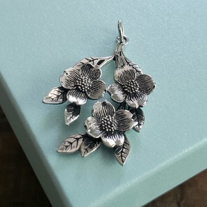 May include: Silver pendant featuring three detailed flower blossoms with leaves. The flowers are stylized and have a textured surface.