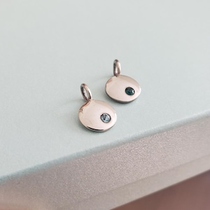 May include: Two small, polished silver pendants. Each pendant has a rounded, oval shape with a small loop at the top for a chain. One pendant features a light blue gemstone, and the other has a dark green gemstone.