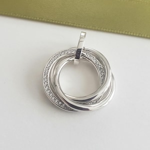 Puede incluir: Colgante plateado con cuatro anillos entrelazados. Un anillo está engastado con pequeñas piedras preciosas transparentes. El colgante tiene un lazo para una cadena. Fondo blanco con una cinta verde.