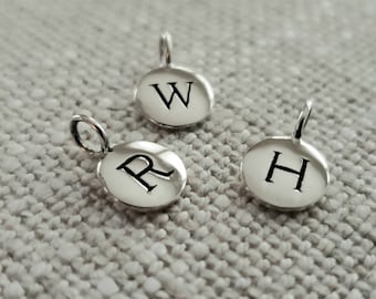 Peut inclure: Trois petits pendentifs argentés de forme circulaire. Chaque pendentif porte une lettre noire différente : "W", "R" et "H". Les pendentifs ont une petite boucle en haut pour les attacher à une chaîne ou un bracelet. Le fond est un tissu gris clair.