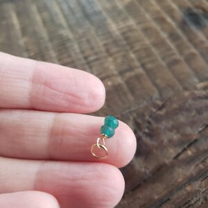 May include: A small, gold-colored jewelry charm with two faceted, emerald-green beads. The charm has a small loop for attaching to a necklace or bracelet. The background is a blurred wooden surface.