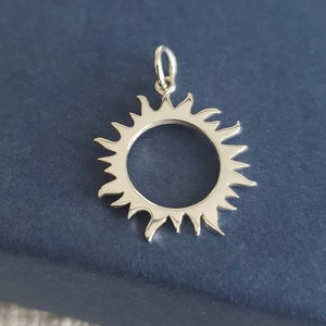 Puede incluir: Un colgante de sol de plata con un diseño circular y un borde texturizado en forma de llama. El colgante está sobre un fondo azul.