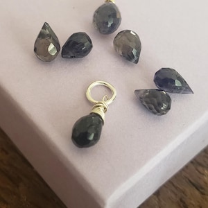 May include: A collection of faceted, teardrop-shaped gemstones in shades of dark blue and gray. One gemstone is wire-wrapped with a silver loop. The gemstones are scattered on a light purple surface, suggesting jewelry components or loose beads.