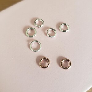 May include: Close-up of silver and rose gold jump rings scattered on a light pink surface. The jump rings are open, ready for use in jewelry making or other crafts. The silver rings are bright and shiny, while the rose gold rings have a warm, metallic hue.
