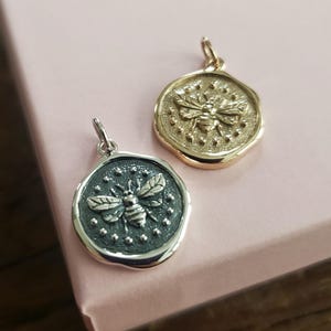 Puede incluir: Dos colgantes de abeja, uno dorado y otro plateado, con un diseño circular texturizado. La abeja está en relieve y detallada.