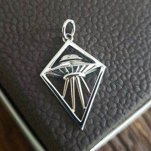 May include: A silver pendant in the shape of a diamond with a silver UFO inside. The UFO has a rounded top and a flat bottom with three legs.